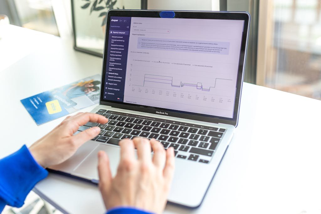 Hands typing on a laptop analyzing business data by a window, showcasing technology in action. Shopify