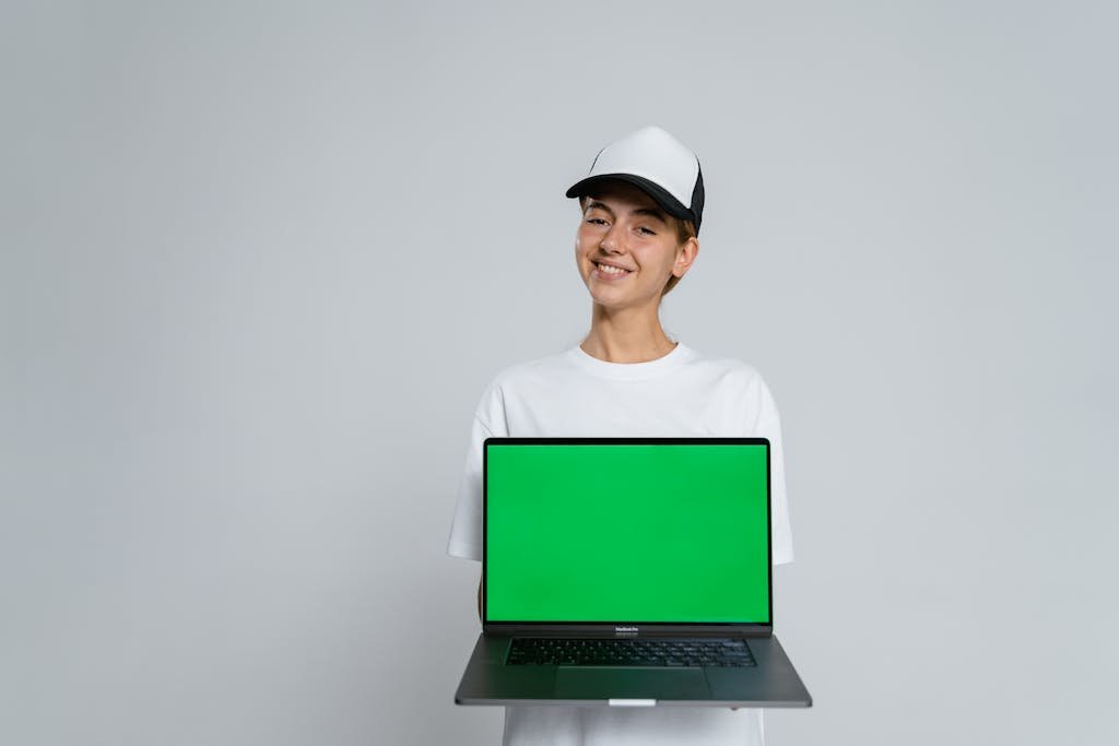 Person in white shirt and cap holding laptop with green screen, ready for mockup.