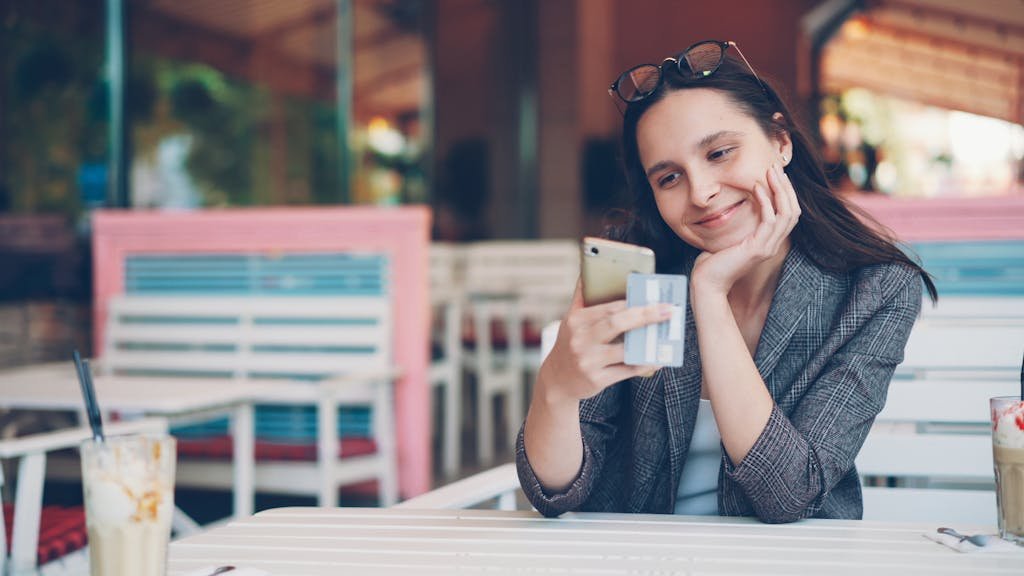 Smiling woman using smartphone and credit card at cafe for online shopping.